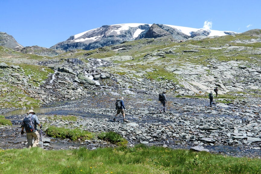 Four people hike across a rocky terrain with patches of grass and a mountain capped with snow in the background under a clear blue sky, experiencing the scenic beauty of Val d'Aosta in Italy.