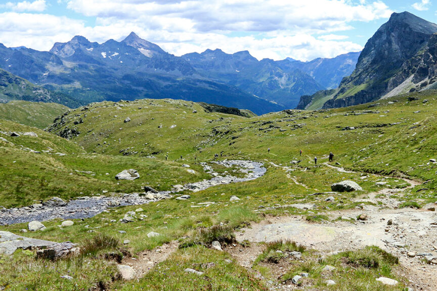 A grassy mountain valley with a trail meandering through it, surrounded by rocky terrain and distant peaks under a partly cloudy sky. Small figures of people are visible on the hiking trail, exploring the beauty of Val d'Aosta in Italy.