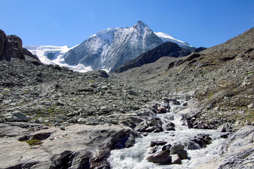 A rocky landscape with a flowing stream in the foreground and a snow-capped mountain under a clear blue sky in the background, reminiscent of the breathtaking vistas encountered on the Haute Route, a famed hiking trail from France to Switzerland.