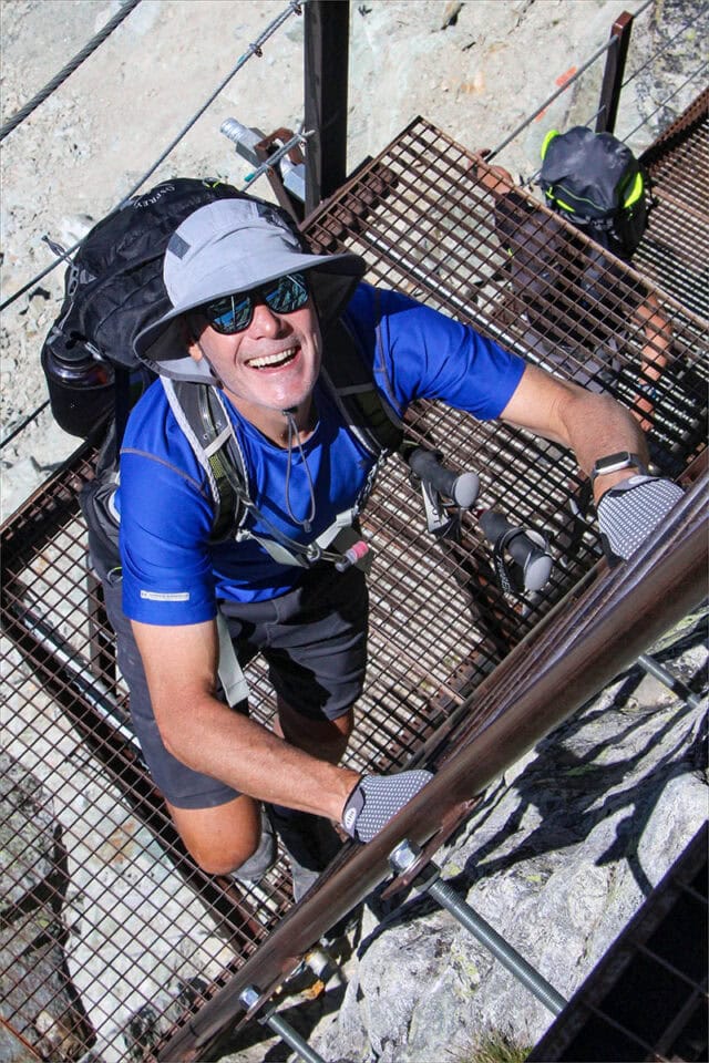 Person in hiking gear smiles while climbing a metal ladder on the rugged Haute Route trail from France to Switzerland, wearing a sun hat, sunglasses, and a backpack.