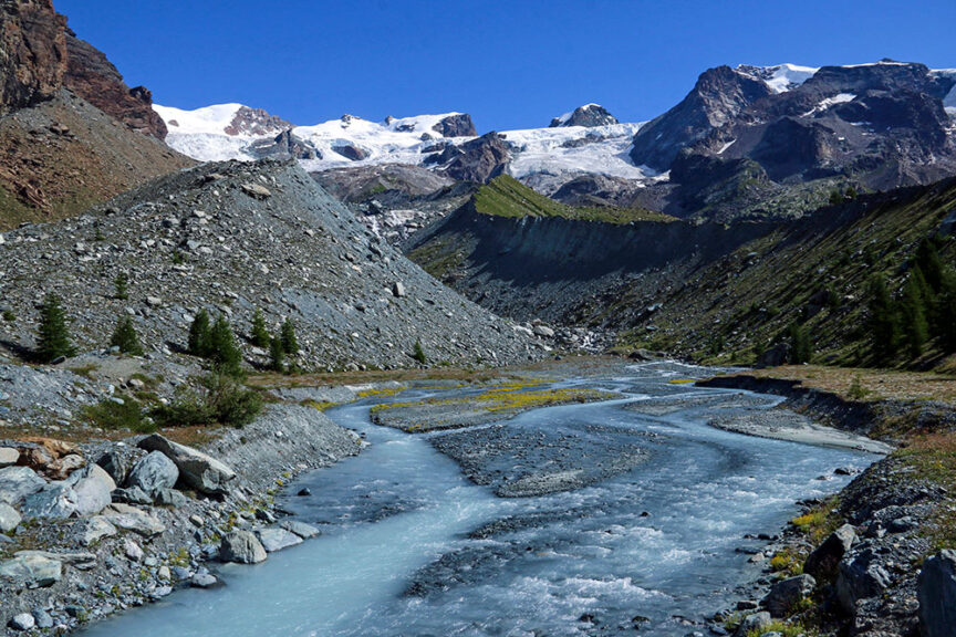 A turquoise river flows through a rocky valley in Val d'Aosta, surrounded by snow-capped mountains under a clear blue sky. Sparse greenery lines the riverbank, offering perfect vistas for hiking enthusiasts exploring this idyllic part of Italy.