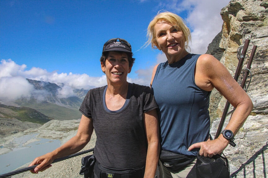 Two women in athletic attire smile at the camera while hiking on the famed Haute Route, with a scenic valley and cloudy sky marking their journey from France to Switzerland.