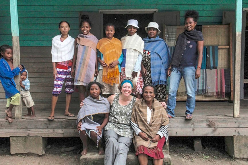 A group of ten people, adults and children, stand and sit on a wooden porch. They are in front of an old wooden building on the island of Madagascar, while some wear traditional shawls, evoking a sense of being lost in time.