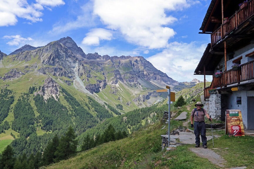 A person with hiking gear stands next to a mountain lodge with a signpost, set against a scenic backdrop of green slopes and rocky peaks under a blue sky with clouds in Val d'Aosta, Italy.