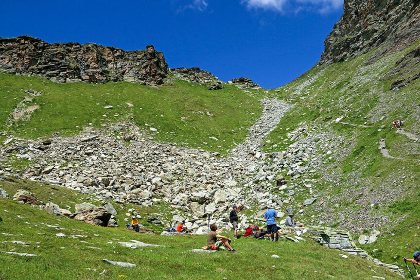 Hikers rest on a grassy alpine hillside in Val d'Aosta with scattered rocks under a clear blue sky, surrounded by rocky mountain slopes, enjoying the picturesque beauty of Italy.