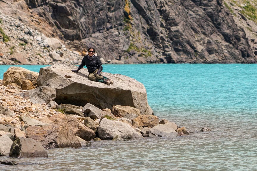 A person sits on a large rock beside a turquoise lake, lost in the wilds of Patagonia, with rocky, mountainous terrain in the background.