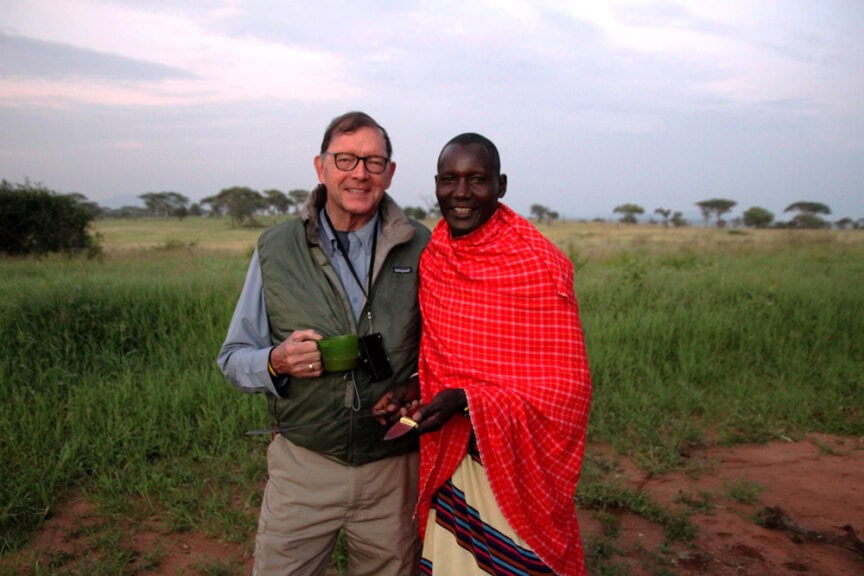 Two individuals stand outdoors smiling; one person holds a green mug, the other wears a red shawl. The background is a grassy landscape in East Africa, extending to the horizon under a cloudy sky.