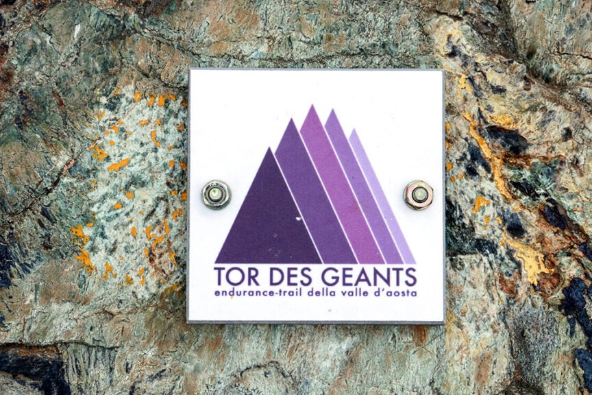 A sign for Tor des Geants, an endurance trail in Val d'Aosta, Italy, is mounted on a rock. The sign features the event name and logo with purple mountain peaks.