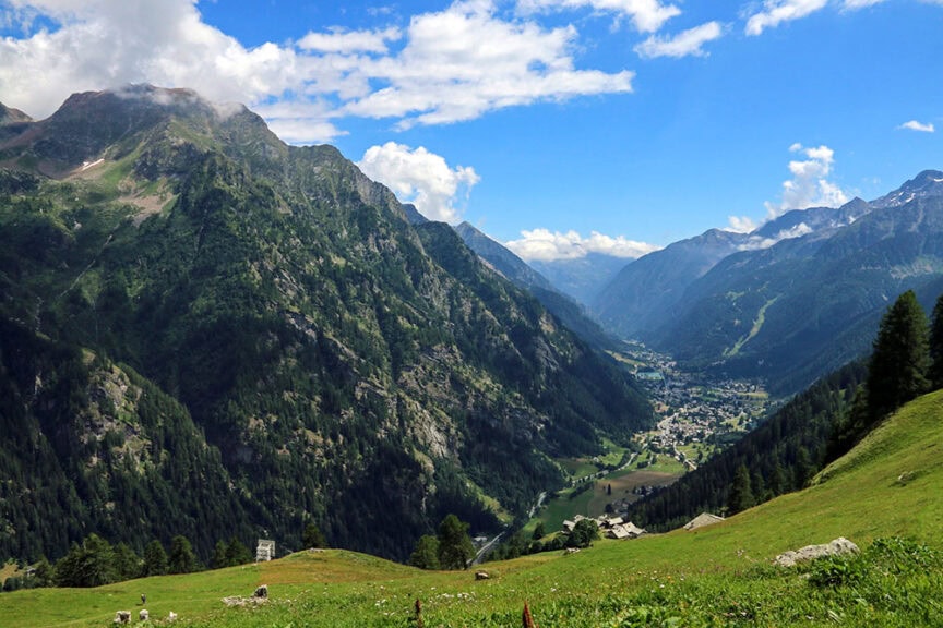 Mountain valley on a sunny day with green slopes, scattered trees, and a small village nestled between the mountains. Blue sky with clouds overhead. Ideal for hiking, this picturesque setting in Val d'Aosta, Italy offers breathtaking views and tranquil surroundings.