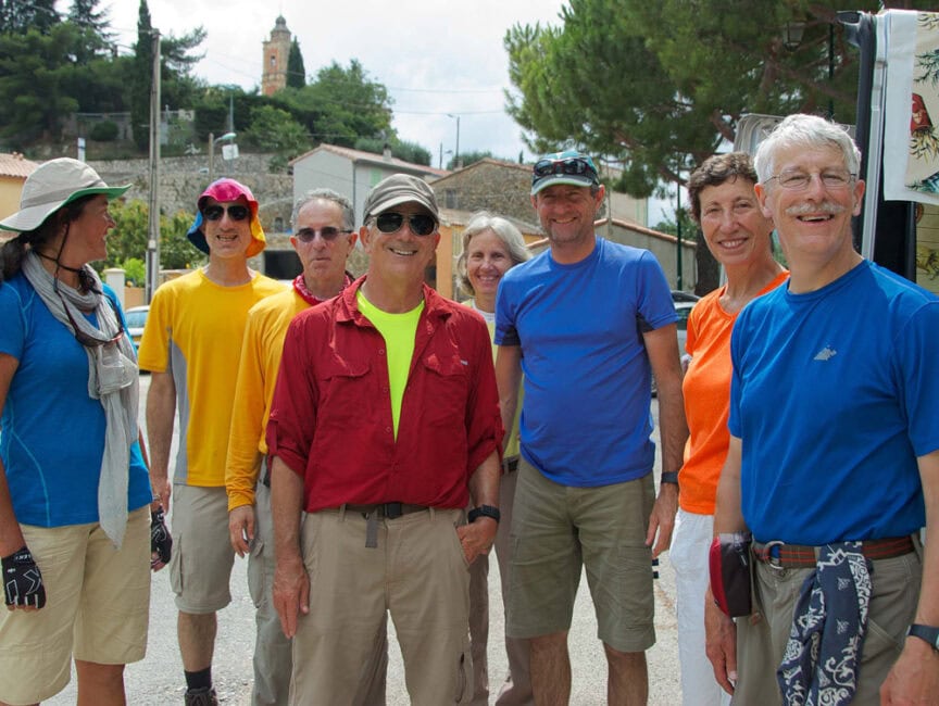 A group of eight people, casually dressed in outdoor clothing, pose for a photo outside on a bright day with towering mountains and trees framing a small building in the background.