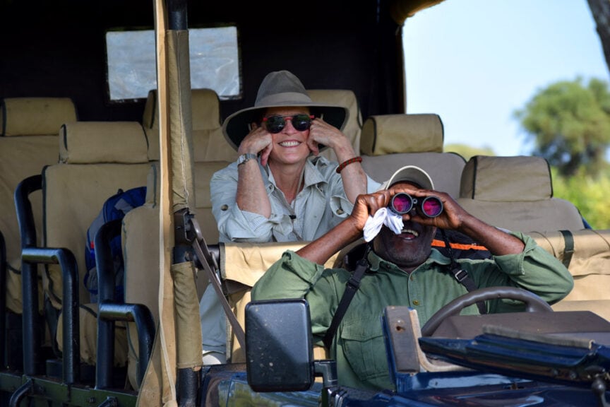 Two people on a safari jeep in East Africa, one wearing a hat and sunglasses, smiling at the camera, and the other exploring the horizon through binoculars with a tissue.