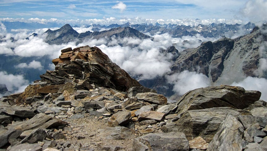 Rocky mountain landscape with clouds floating above the valleys and rugged peaks in the background under a partly cloudy sky, perfect for hiking in the picturesque Val d'Aosta, Italy.