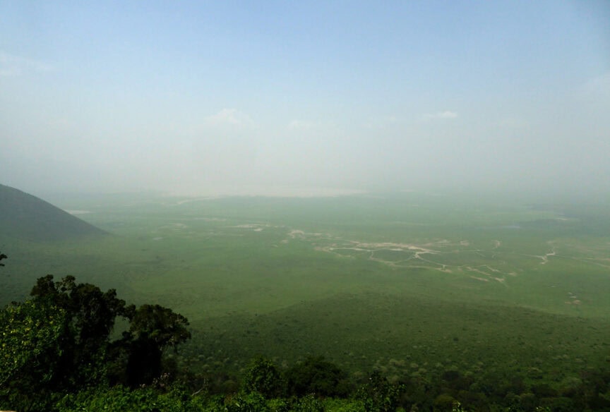 Exploring East Africa, a vast green landscape stretches to the horizon under a hazy sky, with scattered trees in the foreground and a distant flat area.