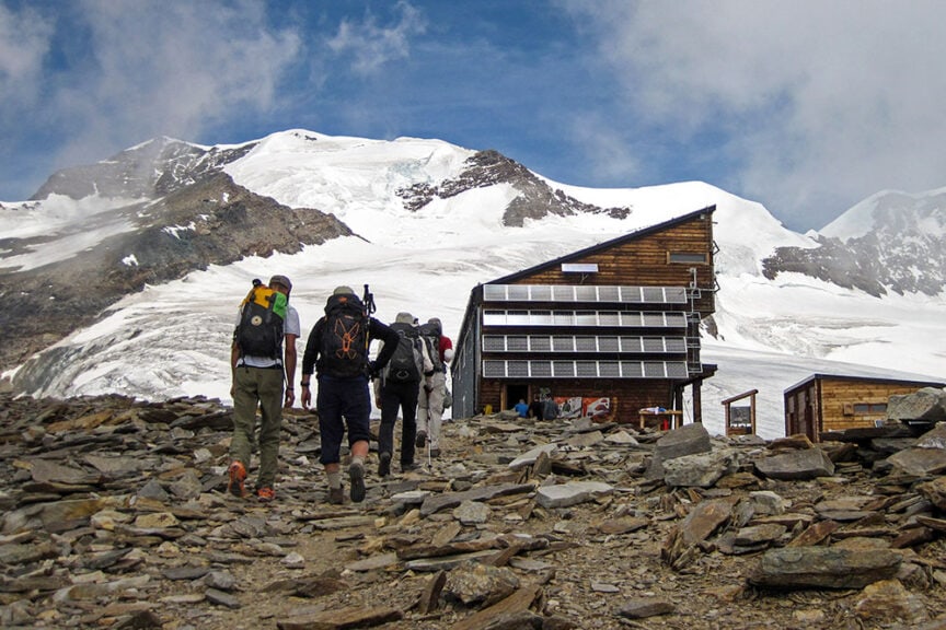 Hikers with backpacks walk towards a wooden mountain lodge with solar panels, set against a snowy alpine backdrop in Italy's picturesque Val d'Aosta, perfect for a memorable hiking adventure.