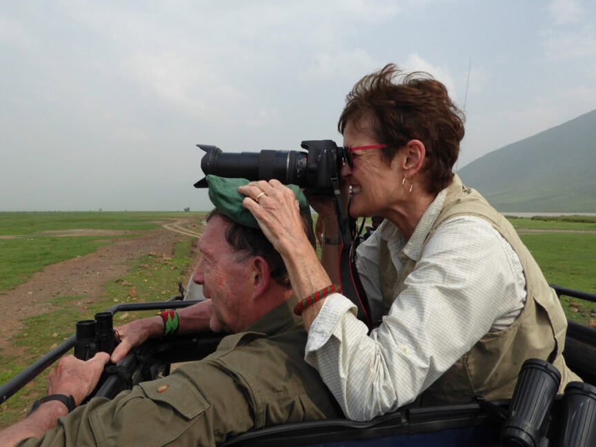 Two people in a safari vehicle are exploring East Africa, looking through binoculars and a camera at wildlife. The woman stands for a better view, holding a telephoto camera, while the man uses binoculars.