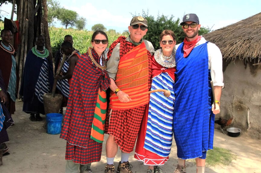 Four people in traditional Maasai attire stand smiling together outside near a thatched-roof structure, embodying the rich culture of East Africa.