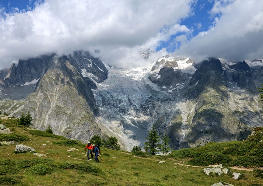 Two hikers traverse a grassy hillside with rocky terrain and patches of greenery in Italy's Val d'Aosta, against a backdrop of towering mountains partially covered by snow and clouds under a blue sky.