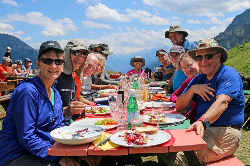 A group of people wearing casual outdoor clothing are sitting at a picnic table in the Val d'Aosta, enjoying a meal together after hiking. The sky over this picturesque part of Italy is clear, and the scene is bright and sunny.