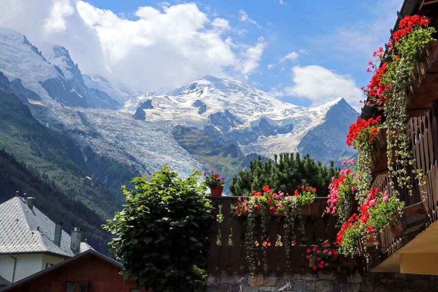 Snow-capped mountains under a blue sky with scattered clouds, seen from a charming village in Val d'Aosta, Italy, with flowers in the foreground. It's a hiking paradise waiting to be explored.
