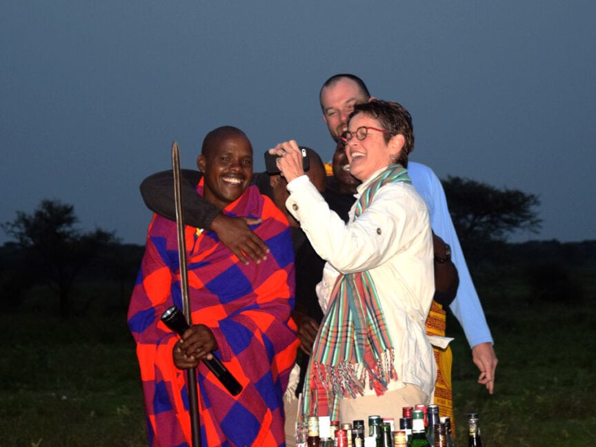 A group of people, one in traditional attire, stand together outdoors, smiling and taking a selfie near a table with various bottles. The background shows a natural, grassy East Africa landscape during dusk.