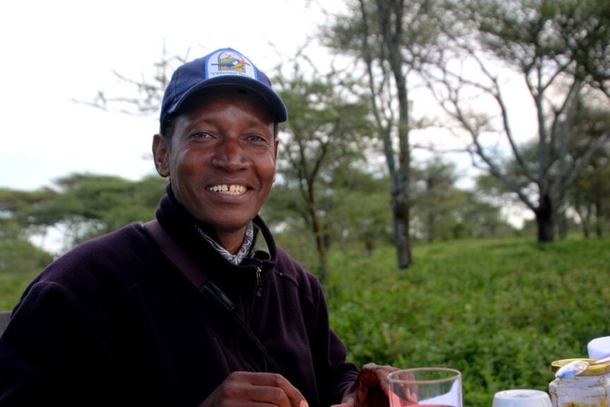 A person wearing a cap smiles while holding a glass outdoors among greenery and trees, embodying the spirit of travel and exploring the scenic landscapes of East Africa.