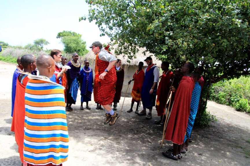 A group of people, some wearing traditional Maasai attire, observe a man in mid-air jumping under a tree in a village setting, capturing the vibrant essence of East Africa.