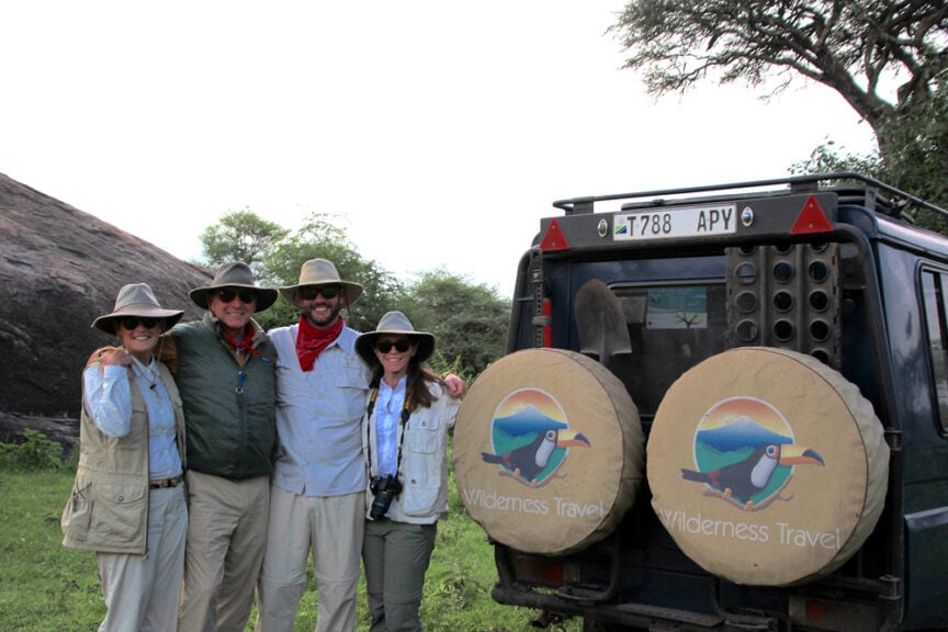 Four people in outdoor gear stand smiling beside a vehicle featuring the Wilderness Travel logo, with spare tire covers depicting a bird. Trees and rocky terrain form the backdrop, capturing the spirit of exploring East Africa.