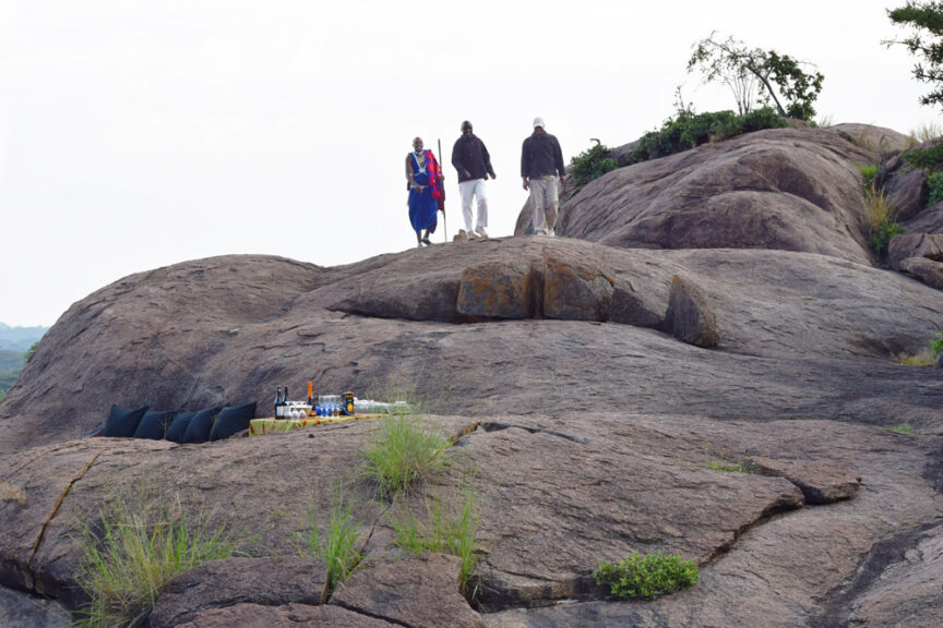 Three people walk on large rocks with a makeshift bar setup on one of the rock surfaces, featuring several bottles and glasses, highlighting their adventurous spirit while exploring East Africa.