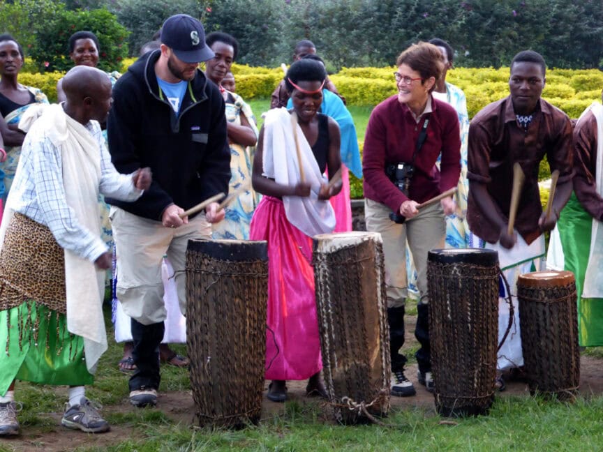 A group of people, including two tourists, are playing large traditional drums outdoors. Amidst the lush greenery, a crowd observes in the background, exploring East Africa's rich cultural heritage.
