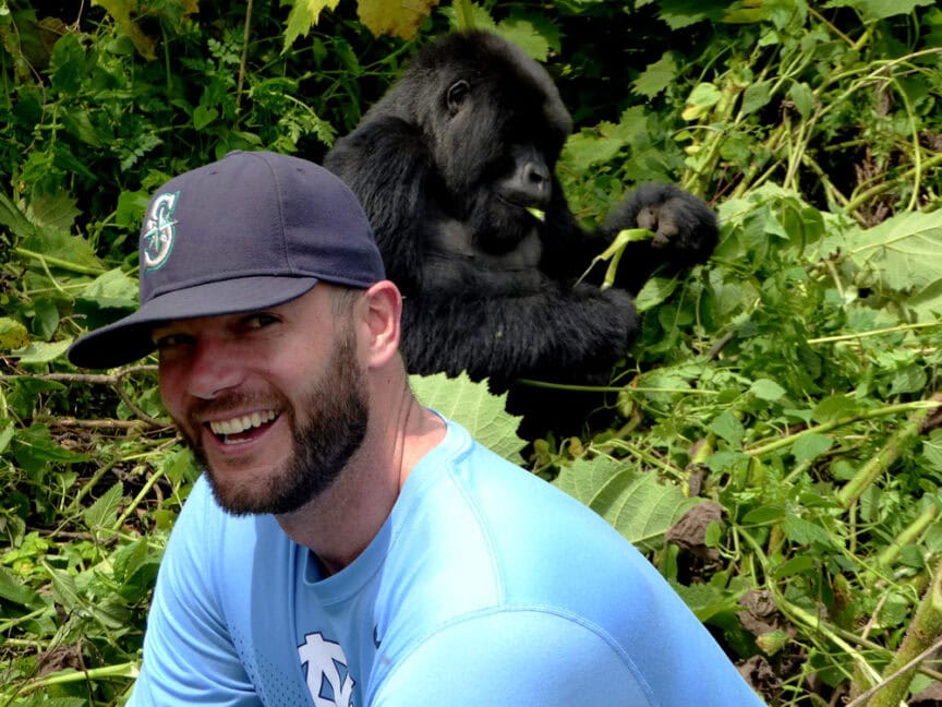 A man wearing a blue shirt and baseball cap smiles at the camera while a gorilla sits behind him, eating vegetation in an East African jungle, embodying the spirit of exploration and travel.