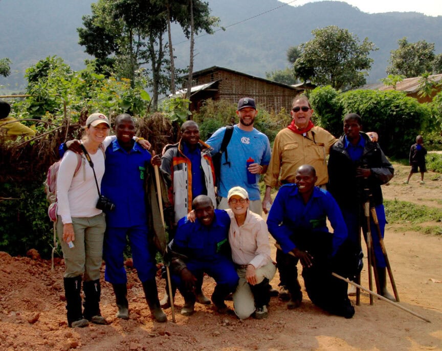 A group of people, including several in blue uniforms and others in casual outdoor clothing, pose together outside in a rural area famous for its tourism, with trees and mountains in the background.