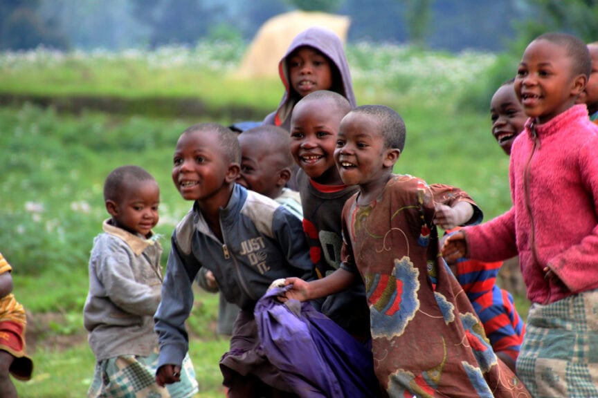 A group of children in colorful clothing run and smile outdoors on a grassy field with trees in the background, exploring the beauty of East Africa.