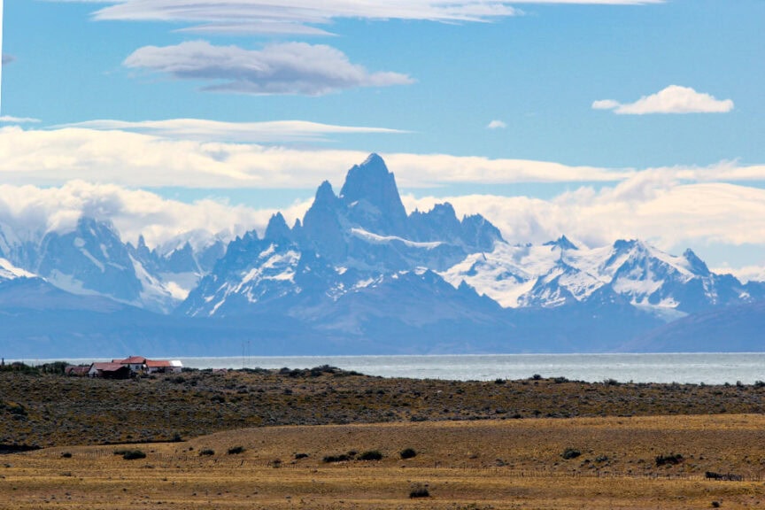 Distant snow-capped mountains with a prominent peak rise above a lake in the wilds of Patagonia. In the foreground, a small cluster of buildings stands on a grassy, barren landscape.