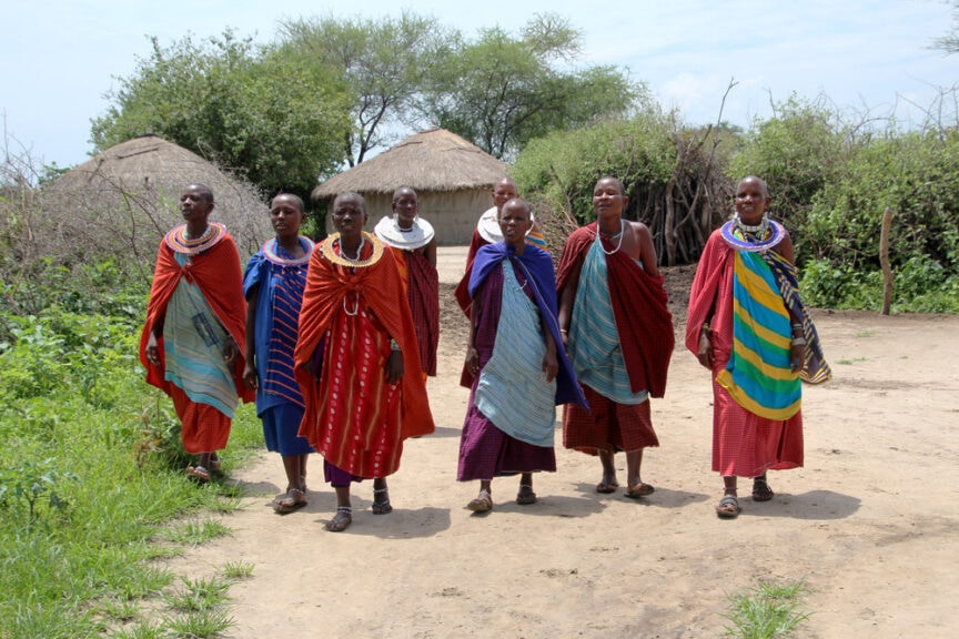 A group of Maasai women in traditional attire, including colorful shukas and beadwork, walk on a dirt path in an East African village with thatched-roof huts and greenery in the background, offering a vivid glimpse into exploring the rich cultural tapestry of the region.