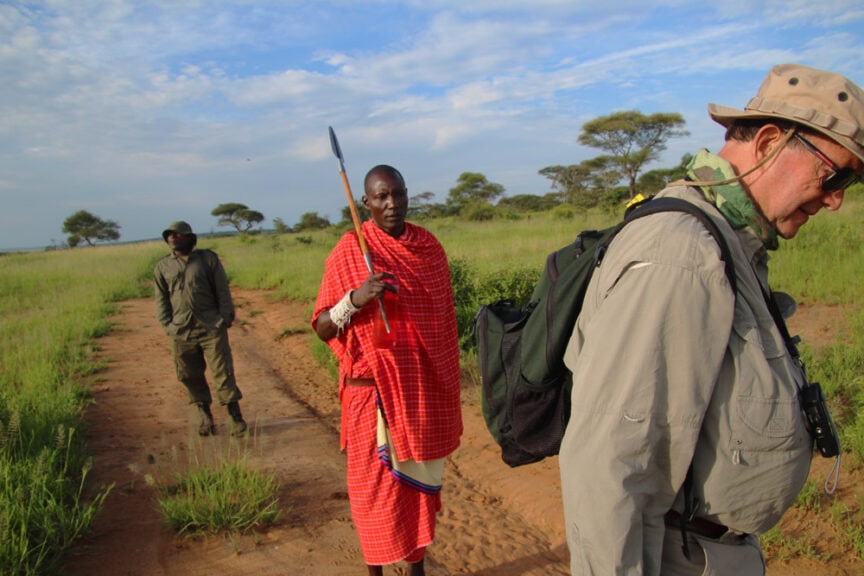Three men walking on a dirt path in a grassy field, perhaps exploring East Africa. One wears a red plaid garment and holds a spear, another is in khaki clothing and hat, and the third is in a green uniform.