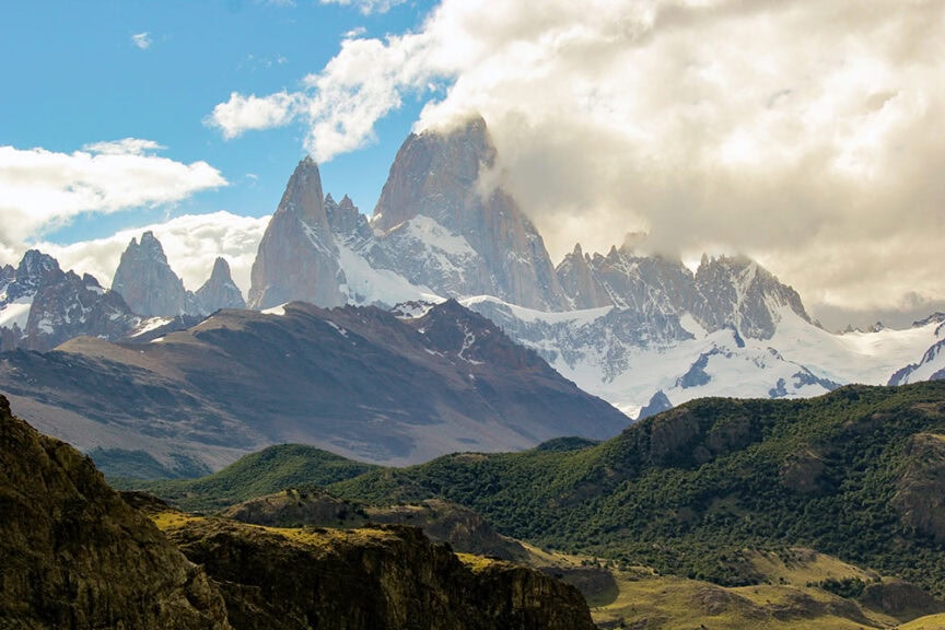 A mountain range with snow-capped peaks partially covered by clouds, surrounded by green and rocky lower hills under a blue sky, capturing the wild beauty of Patagonia.