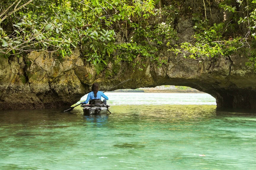 A person kayaking through clear turquoise water, approaching a natural rock archway surrounded by lush green vegetation in the stunning Rock Islands of Palau, often referred to as the Underwater Serengeti.