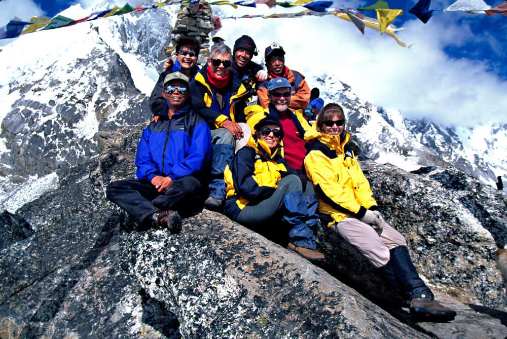 A group of climbers in colorful jackets pose together on a rocky mountain slope with snowy peaks and prayer flags in the background, capturing a memorable moment for their Nepal photobook.