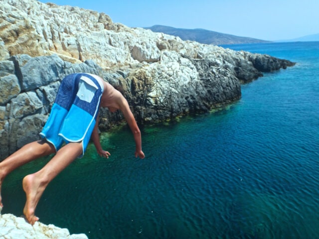 A person in blue swim trunks is diving off a rocky cliff into a calm, clear blue sea, capturing the vibrant colors of the Aegean with rocky cliffs and hills in the background.