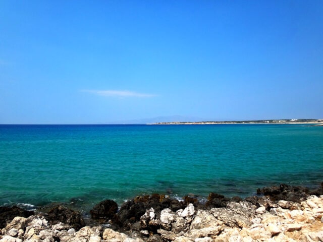 A view of a rocky shoreline with the vibrant colors of the Aegean Sea's clear blue-green water extending to the horizon, under a bright blue sky.