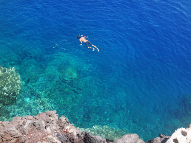 A person snorkeling in the clear turquoise waters of the Aegean, near a rocky shoreline vibrant with colors.