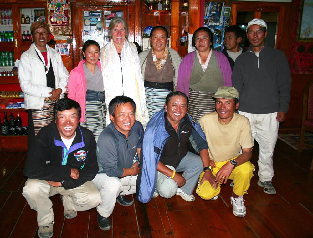 A group of men and women, resembling a close-knit family, stand and kneel together in a cozy, wooden-floored room lined with shelves holding various items. Their joyous expressions hint at shared memories that belong in a treasured photobook from Nepal.