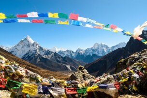 Colorful prayer flags are strung across a mountainous landscape with clear blue skies and snow-capped peaks in the background, capturing the essence of a trek in Nepal. Remember to pack your trekking essentials for this awe-inspiring journey.