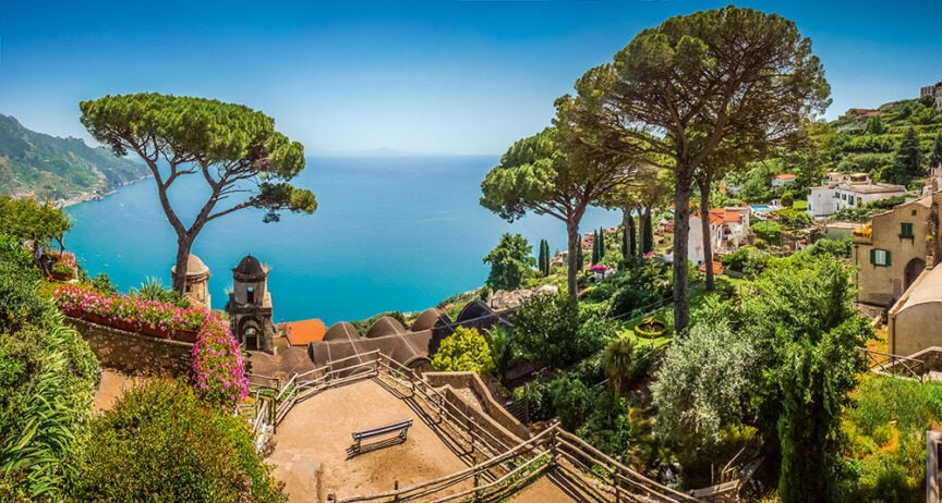 Coastal townscape with lush greenery, tall trees, and buildings overlooking a deep blue sea. There are several benches along the pathway, and vibrant flowers border some areas, making it a picturesque spot for travelers exploring Europe.