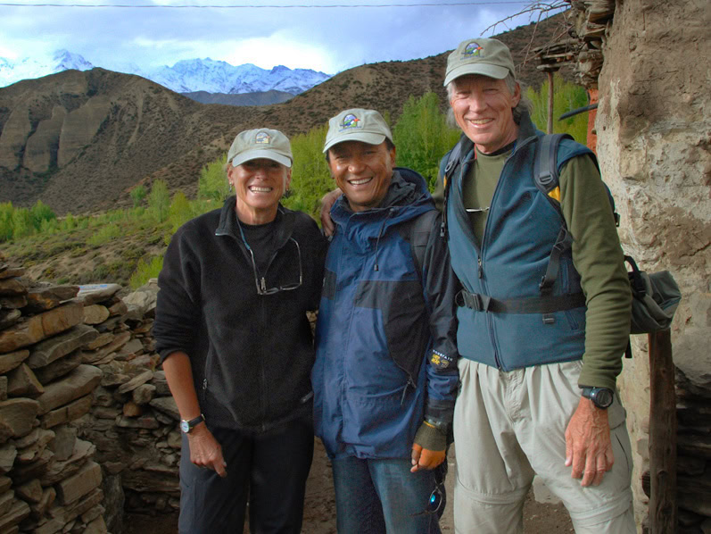 Three hikers, perhaps a family, wearing caps and outdoor gear pose for a photo with the stunning mountainous landscape of Nepal in the background. Perfect for a photobook memory!
