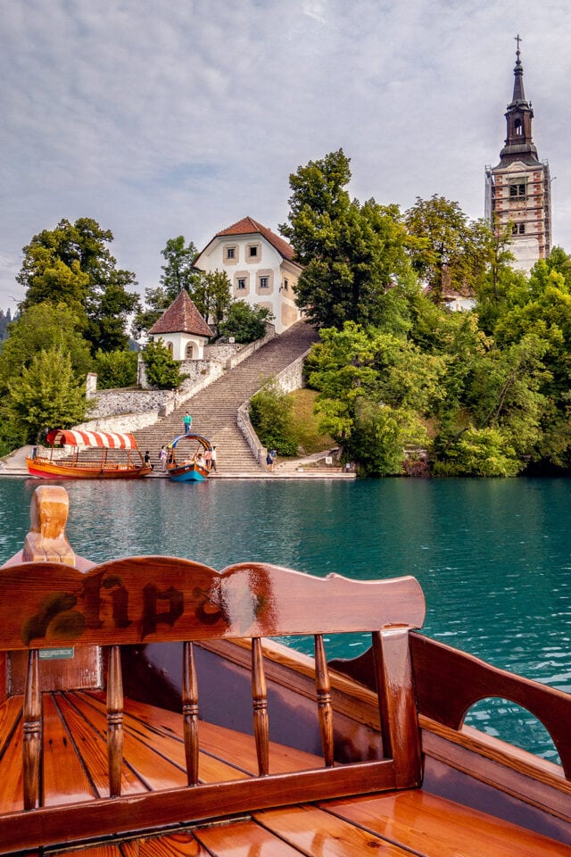 Wooden boat pointing towards an island with a church and other buildings surrounded by greenery. Stairs lead from the waterfront to the church. People walking and boats docked along the shore, perfect for a Slovenia hiking adventure.