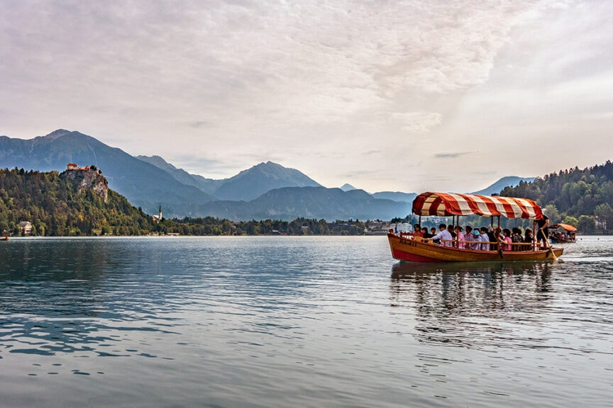 A boat with a striped canopy carries a group of people on Slovenia's picturesque lake, surrounded by rugged mountains, with a castle visible on a distant cliff.