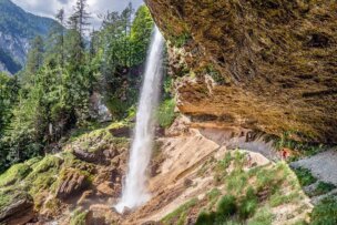 A waterfall cascades from a rocky cliff surrounded by green forest, providing a perfect setting for wild hiking. People walk on a path behind the waterfall, immersing themselves in Slovenia's rugged adventure.