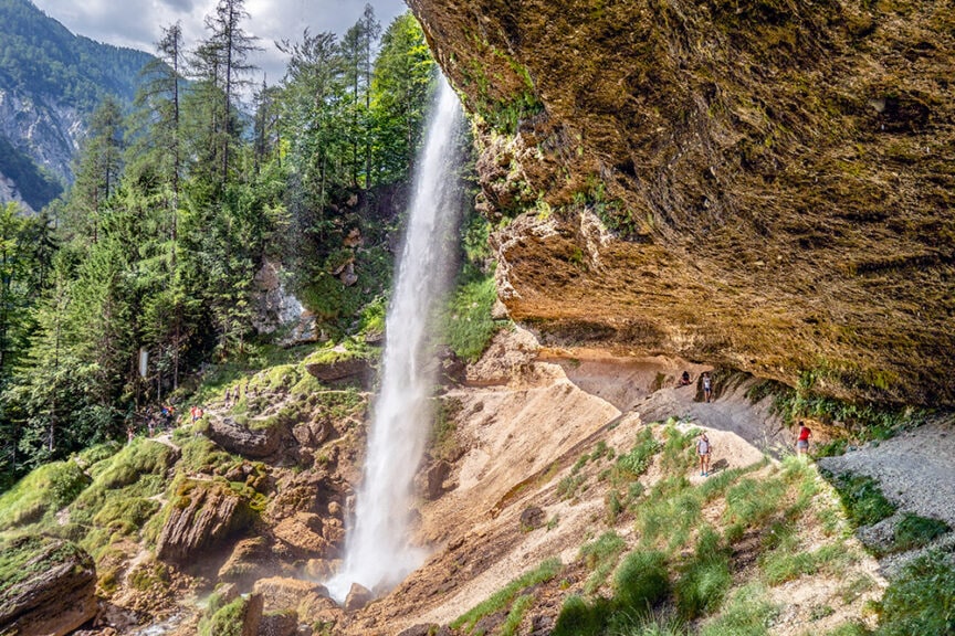 A waterfall cascades from a rocky cliff surrounded by green forest, providing a perfect setting for wild hiking. People walk on a path behind the waterfall, immersing themselves in Slovenia's rugged adventure.