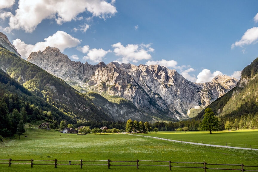A picturesque mountain range rises behind a rugged green valley with scattered houses and trees under a partly cloudy blue sky, setting the stage for a wild hiking adventure.